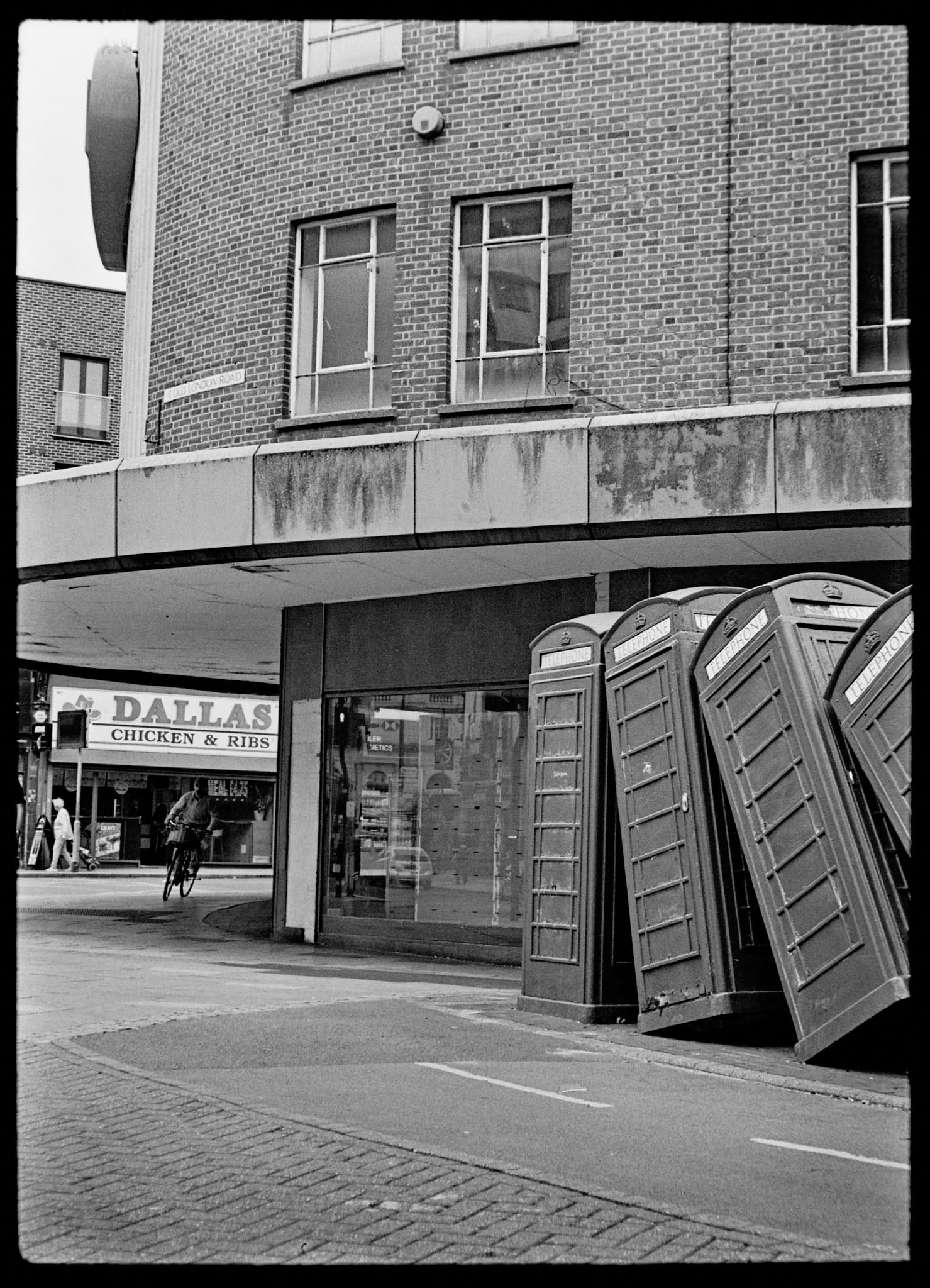 A digital scan of a black-and-white film negative of 'Out of Order' in Kingston upon Thames, a sculpture by David Mach depicting several red telephone boxes falling like dominoes.